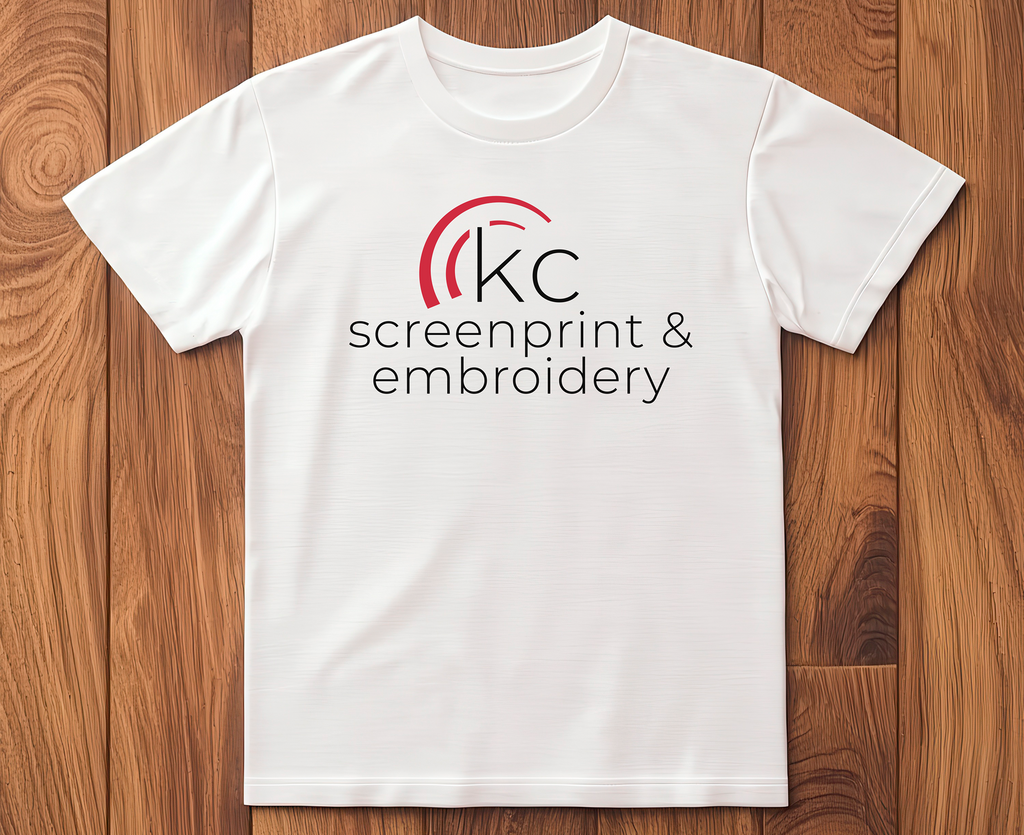 White t-shirt with 'kc screenprint & embroidery' logo on a wooden surface