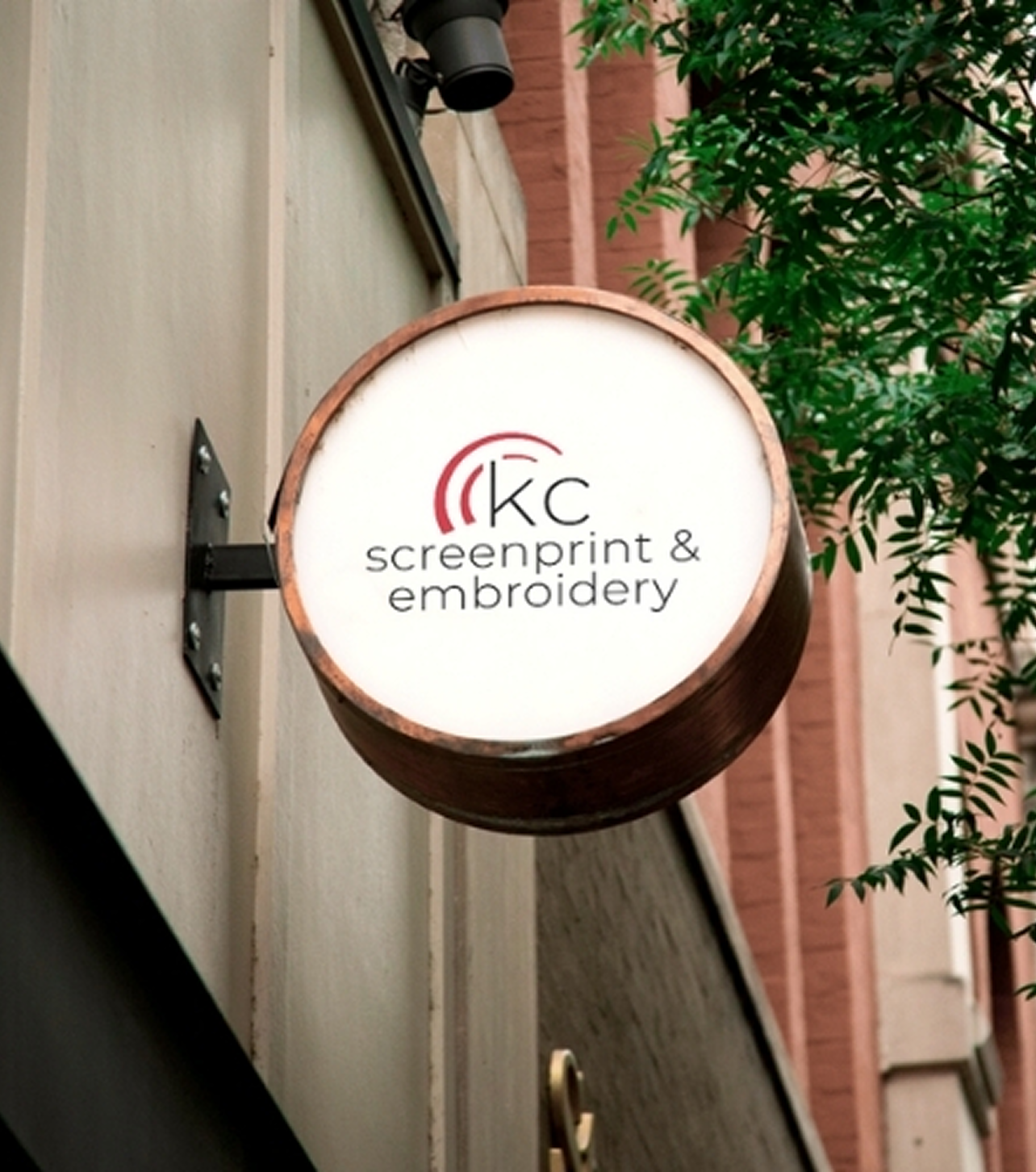 Round signboard with 'KC screenprint & embroidery' logo on a building exterior.